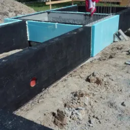 Fundamenty domu w trakcie budowy: czarna izolacja przeciwwilgociowa, niebieski styropian, wystające pręty zbrojeniowe, czerwona kurtka na prętach, widoczna rura kanalizacyjna w ścianie fundamentu.