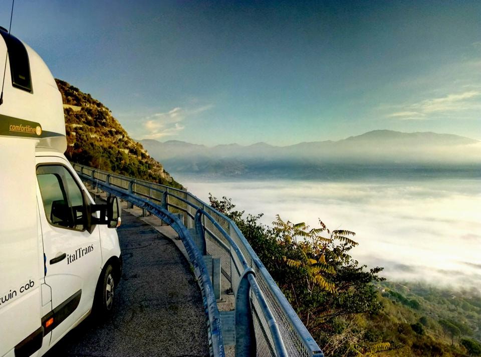 Biały van firmy ItalTrans z widokiem na górzysty krajobraz spowity mgłą, sfotografowany z krętej drogi z barierkami.