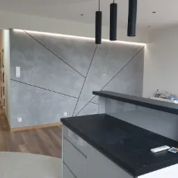 Szara ściana z geometrycznym wzorem i podświetleniem LED, widok z perspektywy baru kuchennego z czarnym blatem.