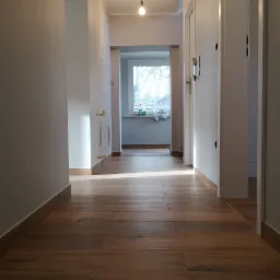Długi korytarz z drewnopodobną podłogą z płytek, świeżo pomalowane białe ściany, widok na okno z bliska