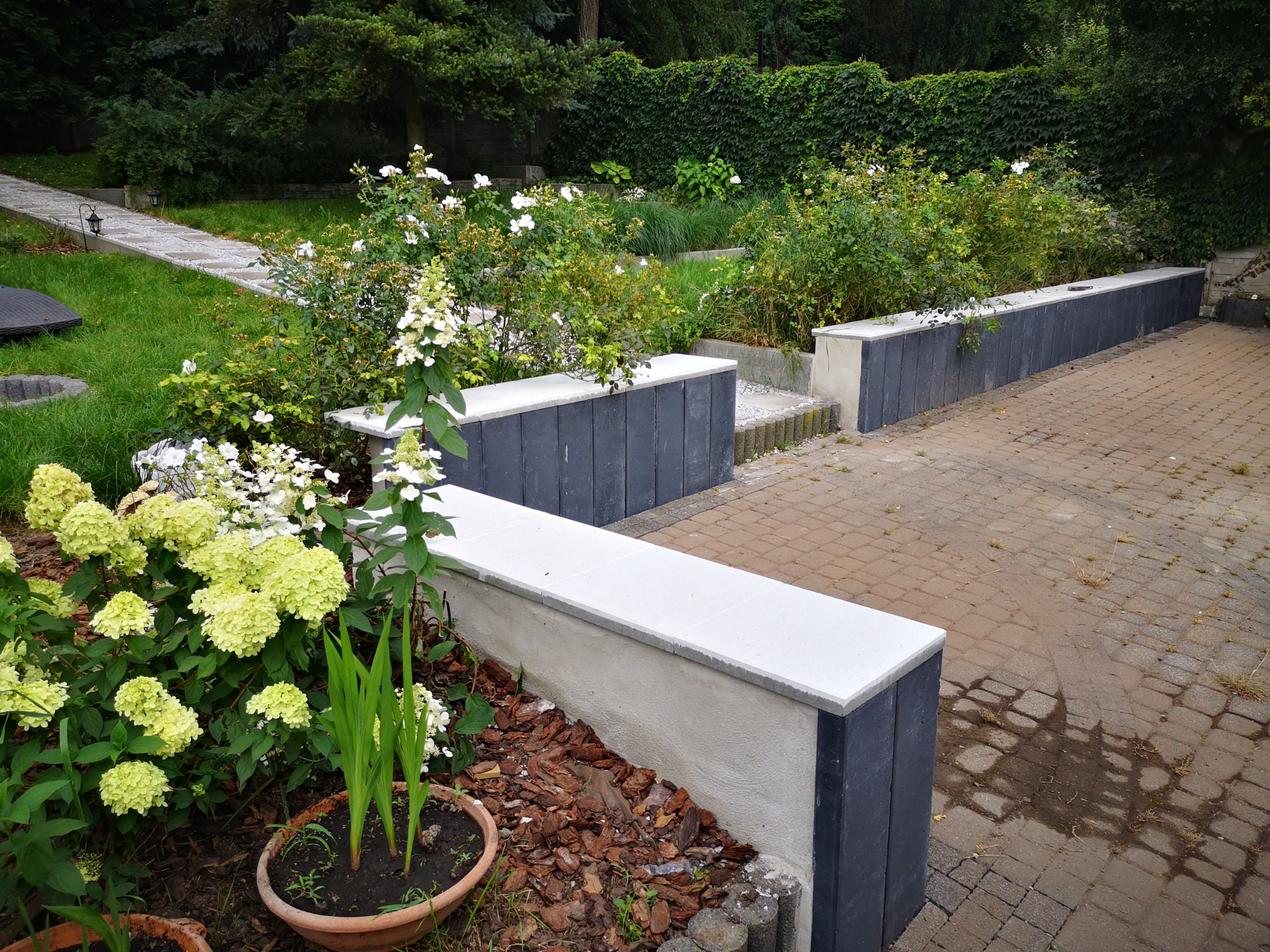 Murowana rabata z szarymi panelami i białym blatem, otoczona kwitnącymi hortensjami i brukowanym chodnikiem w zielonym ogrodzie.