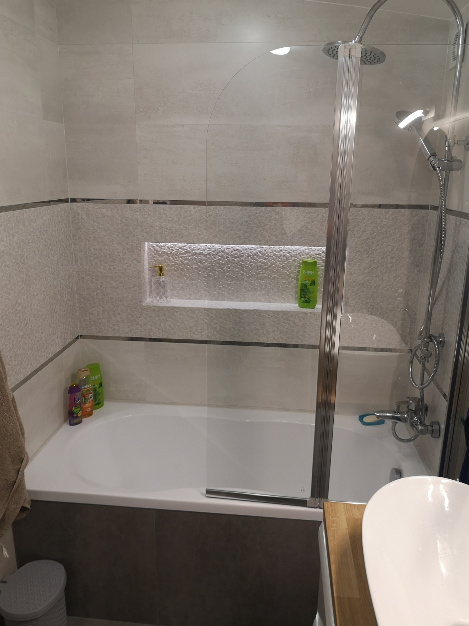 Wykończona łazienka z wanną zabudowaną szarą mozaiką, białe płytki na ścianach z wnęką oświetloną LED, szklany parawan nawannowy, widoczny fragment umywalki.