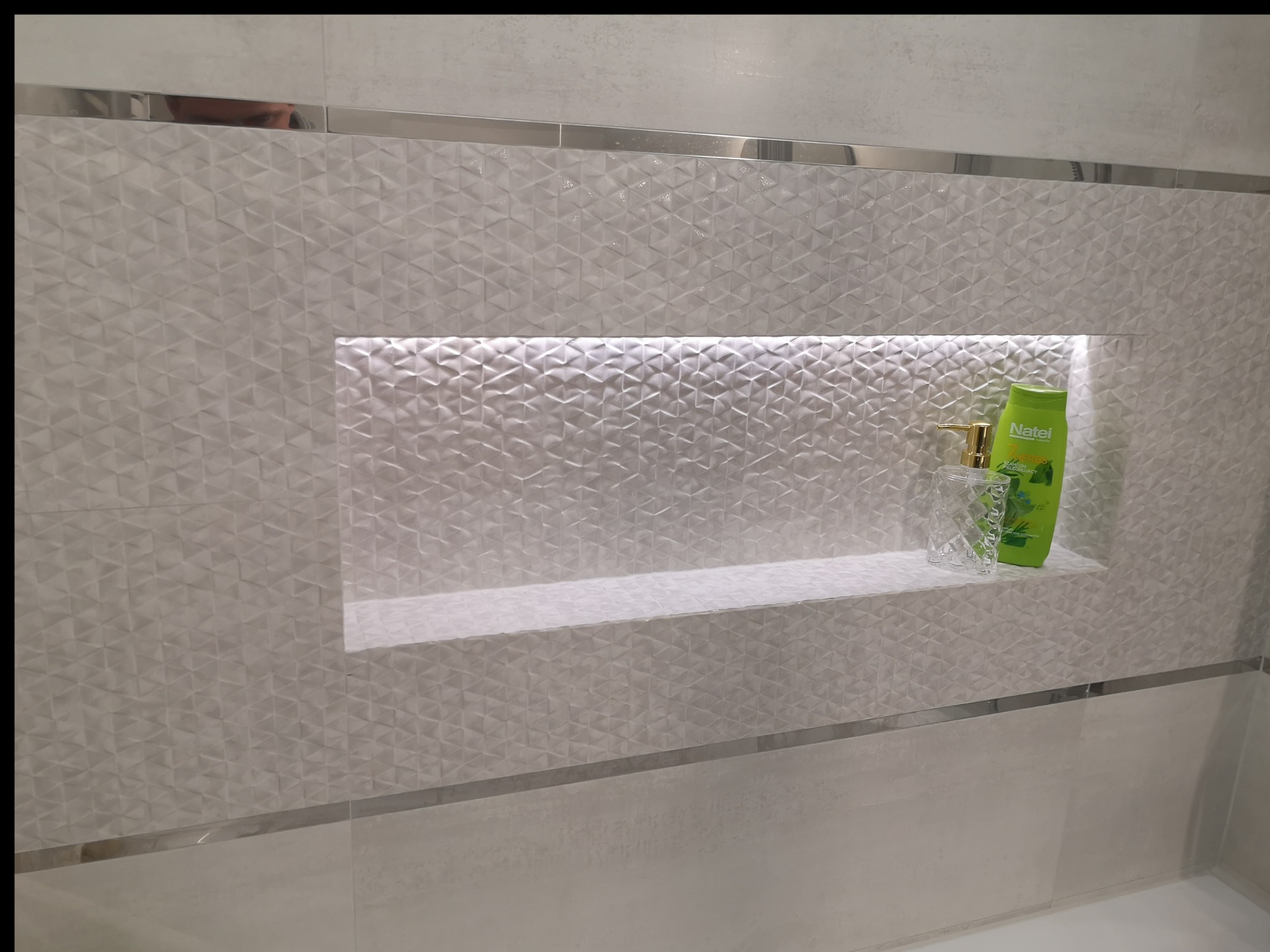 Biała ściana w łazience wyłożona trójwymiarowymi płytkami z wnęką oświetloną od góry, na której stoją mydło w płynie i szampon.