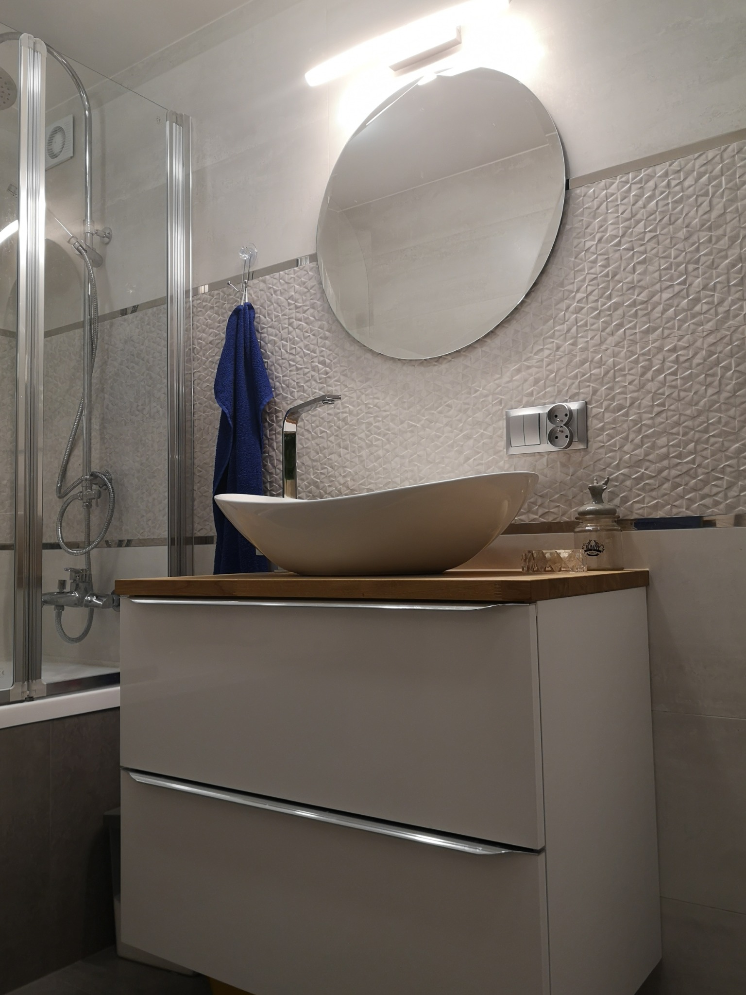 Nowoczesna łazienka z owalną umywalką nablatową, baterią w kolorze chromu, okrągłym lustrem, płytkami z trójwymiarowym wzorem i kabiną prysznicową ze szklanymi drzwiami.