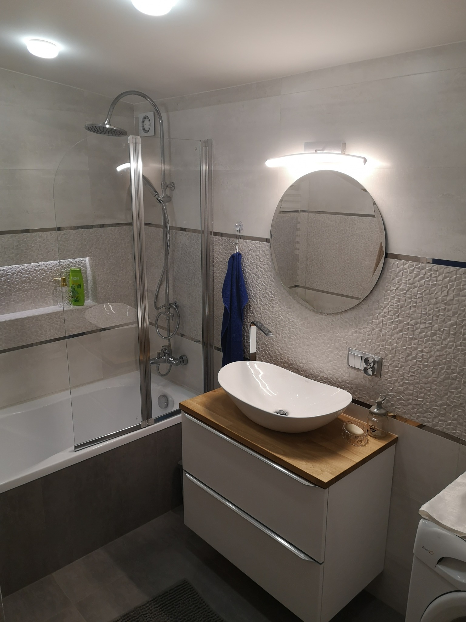 Nowoczesna łazienka z owalną umywalką nablatową na drewnianym blacie, kabiną prysznicową ze szklanymi drzwiami oraz okrągłym lustrem z podświetleniem LED.