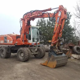 Pomarańczowa koparka kołowa Atlas 1404 w Szamotułach, zaparkowana na placu budowy z widocznym wywrotką w tle, otoczona drzewami i stertą opon oraz metalowych elementów.