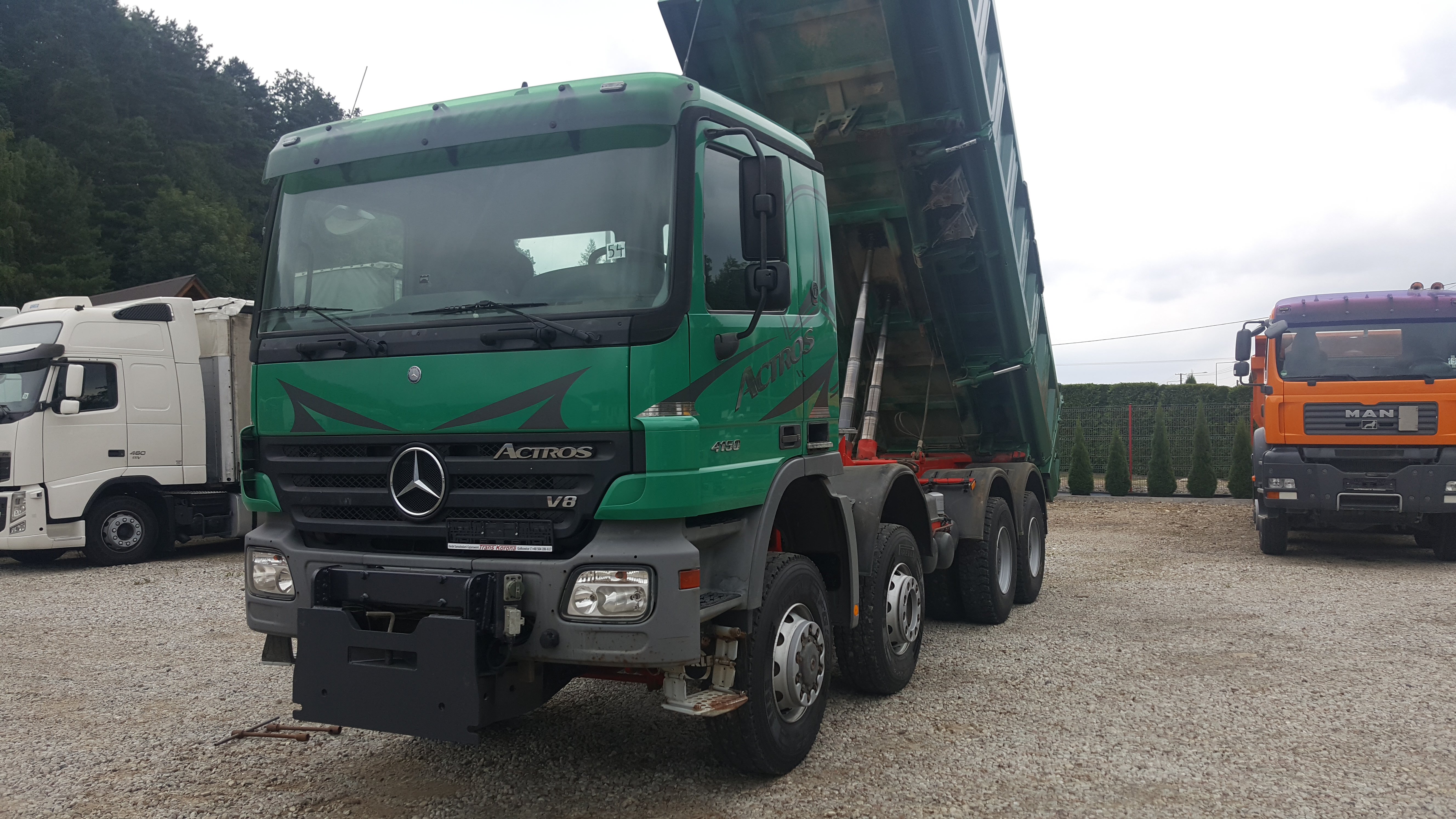 Zielona wywrotka Mercedes Actros 4150 z podniesioną skrzynią ładunkową na żwirowym placu, widoczne inne ciężarówki w tle.
