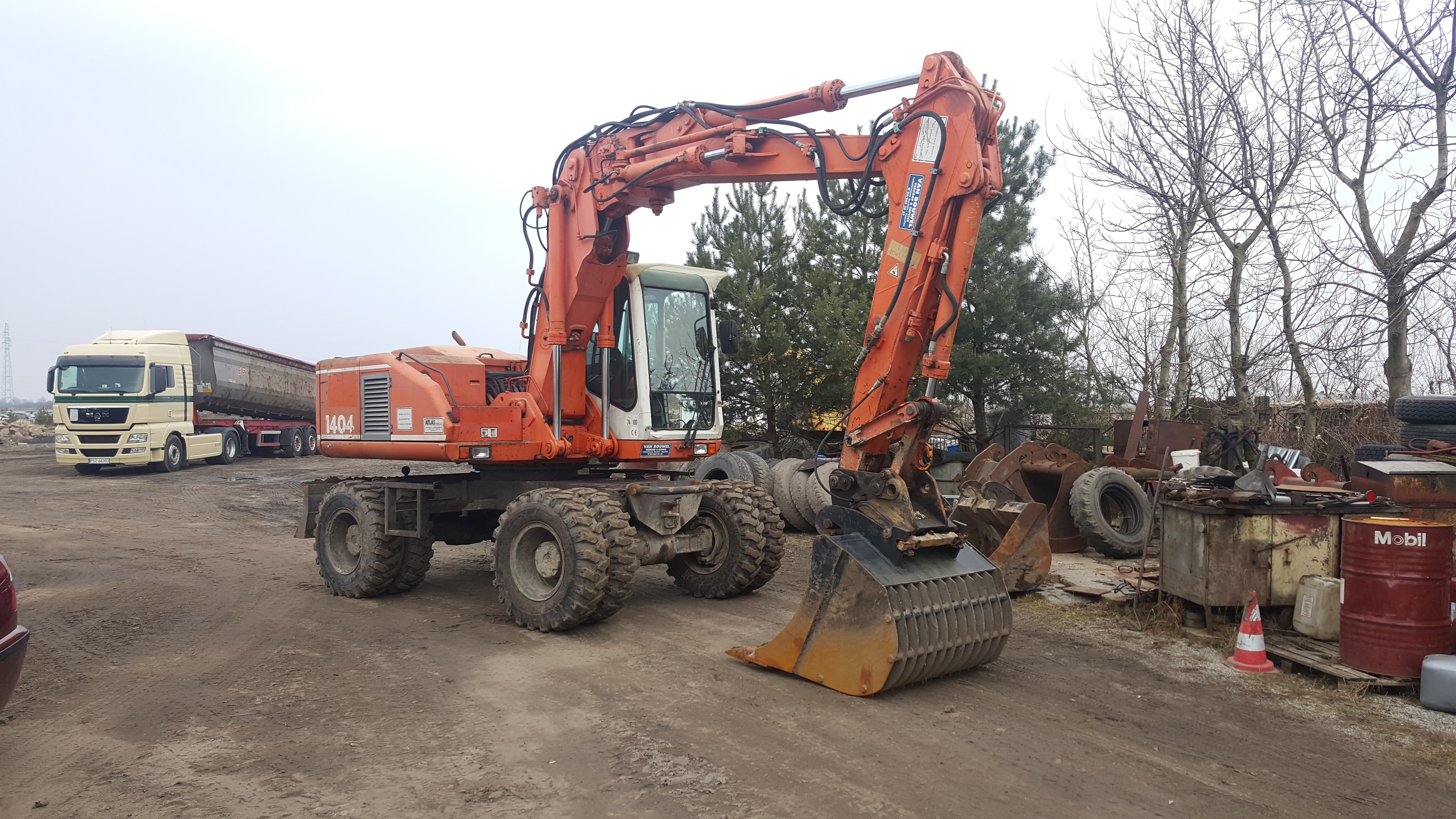Pomarańczowa koparka kołowa Atlas 1404 w Szamotułach, zaparkowana na placu budowy z widocznym wywrotką w tle, otoczona drzewami i stertą opon oraz metalowych elementów.