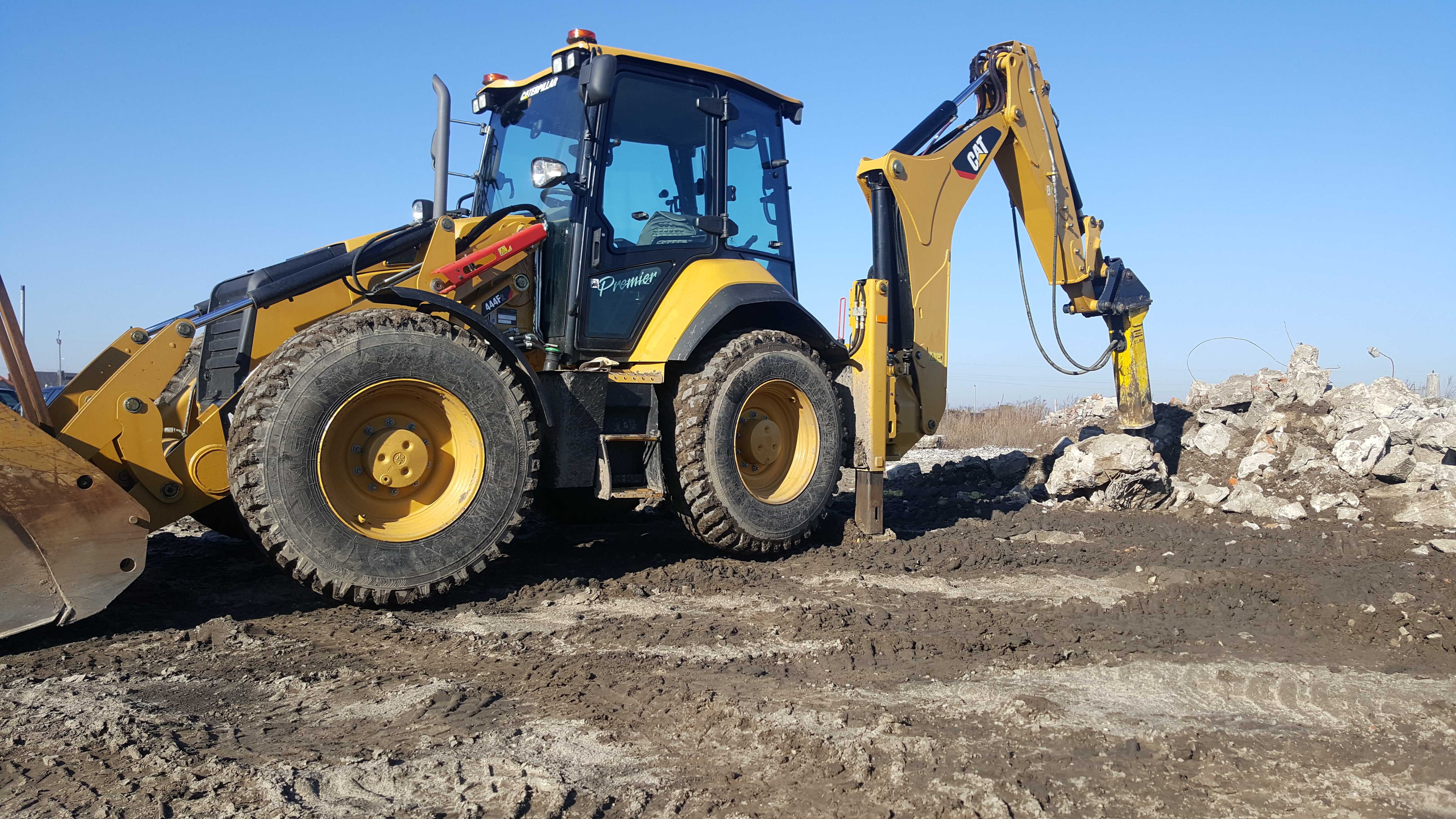 Żółta koparko-ładowarka Caterpillar z hydraulicznym młotem pneumatycznym rozbija gruz na placu budowy w słoneczny dzień.