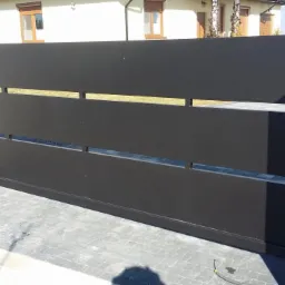 Nowoczesna, przesuwna brama wjazdowa w kolorze antracytowym z poziomymi szczelinami, osadzona między dwoma białymi słupkami na tle brukowanej nawierzchni i domu.