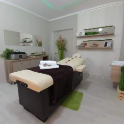 Jasny gabinet z beżowym łóżkiem do zabiegów, brązowym pledem, białymi ręcznikami i płatkami róż, w tle szafka z podgrzewaczem i dekoracjami.