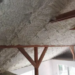 Poddasze z drewnianą konstrukcją pokryte grubą warstwą jasnej piany izolacyjnej, widoczne metalowe elementy mocujące oraz fragment okna w tle.