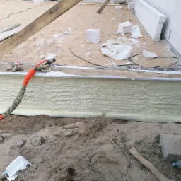 Świeżo natryśnięta, jasna izolacja piankowa na fundamencie budynku w trakcie budowy. Widoczne narzędzie do natrysku, resztki materiałów budowlanych i piasek.