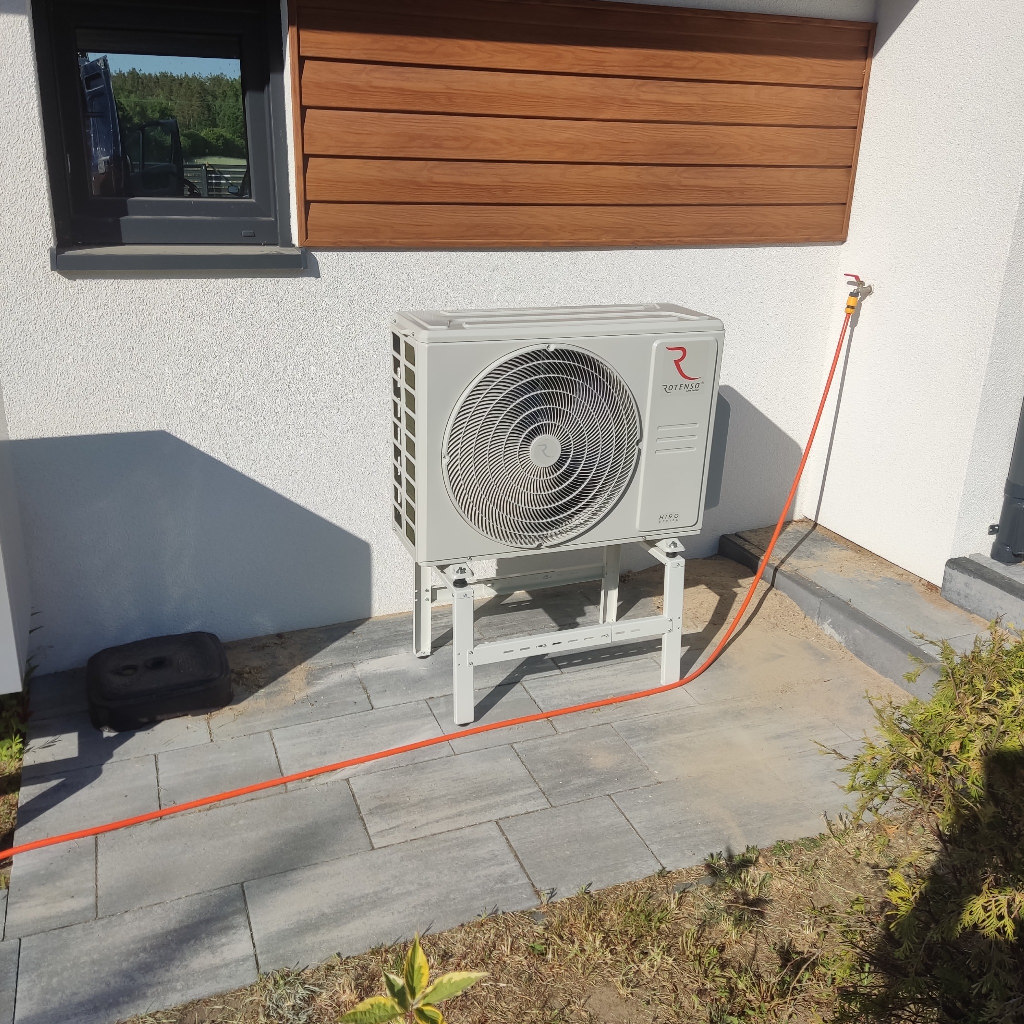 Zewnętrzna jednostka klimatyzacji Rotenso HIRO Series zamontowana na białym stelażu przy ścianie budynku w Starogardzie Gdańskim. Widoczny kran z podłączonym pomarańczowym wężem.