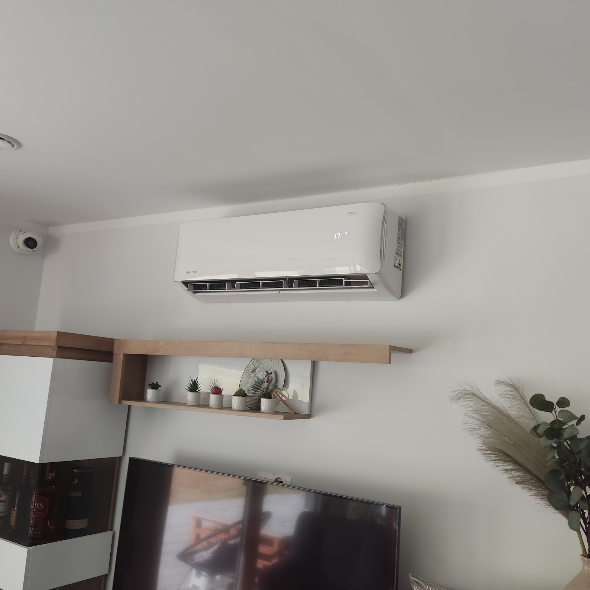 Biała jednostka klimatyzacji ściennej marki Rotenso, z wyświetlaczem temperatury 17 stopni Celsjusza, zamontowana w nowoczesnym salonie z drewnianą półką dekoracyjną i telewizorem.