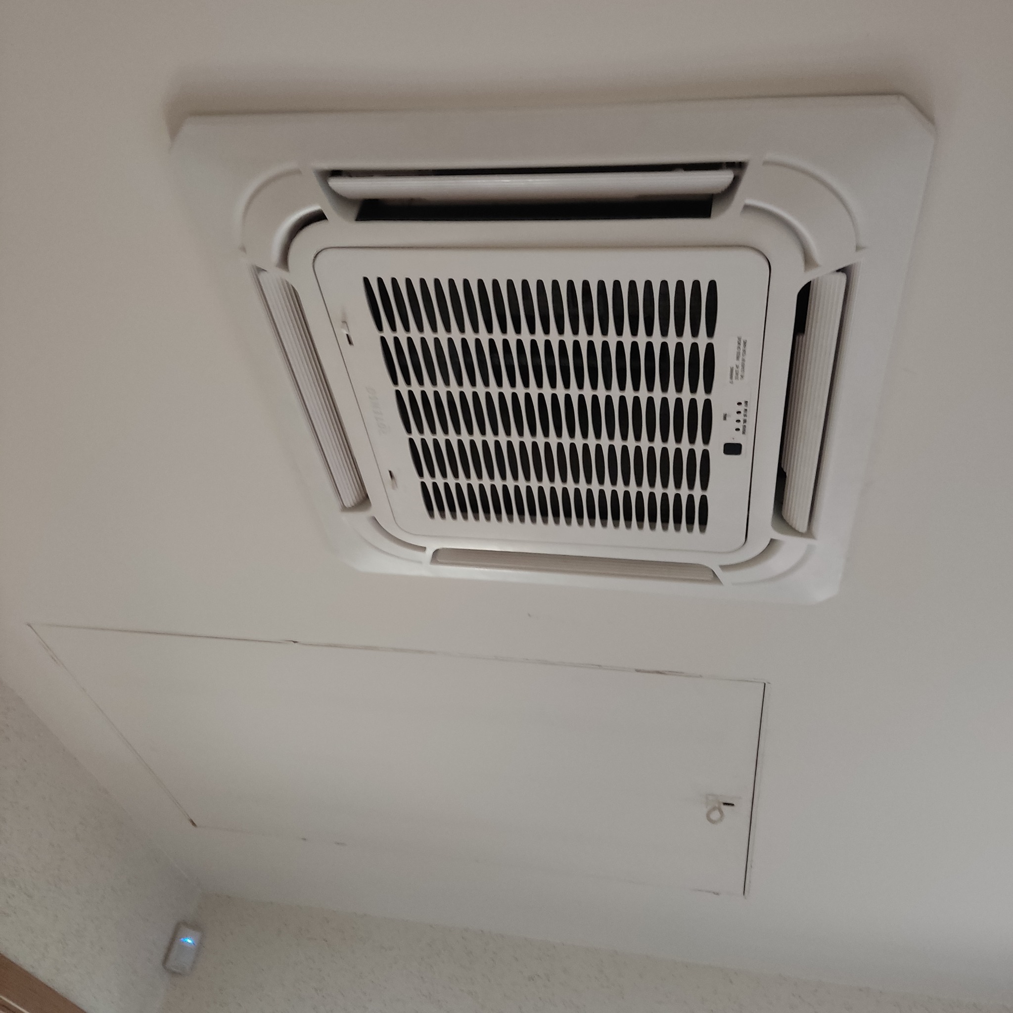 Biała, sufitowa kaseta klimatyzacyjna zamontowana w pomieszczeniu z dostępem do instalacji przez klapę rewizyjną.