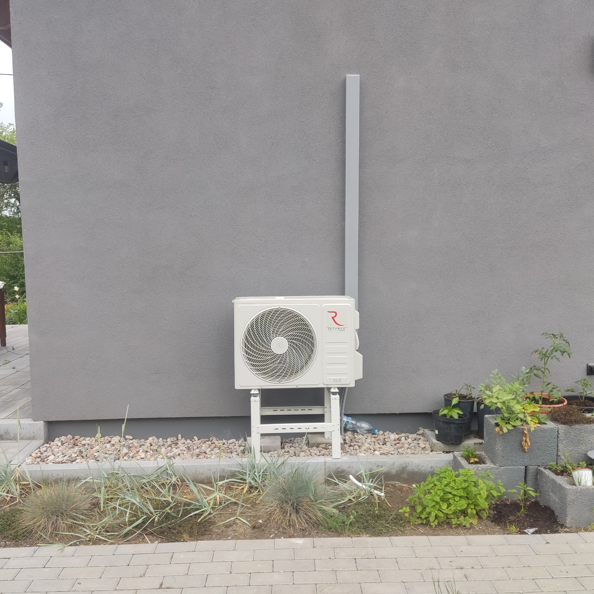 Biała jednostka zewnętrzna klimatyzacji marki Rotenso zamontowana na białym stelażu przy szarej ścianie budynku, widoczna instalacja odprowadzająca na górę, obok dekoracyjne kamienie i rośliny...