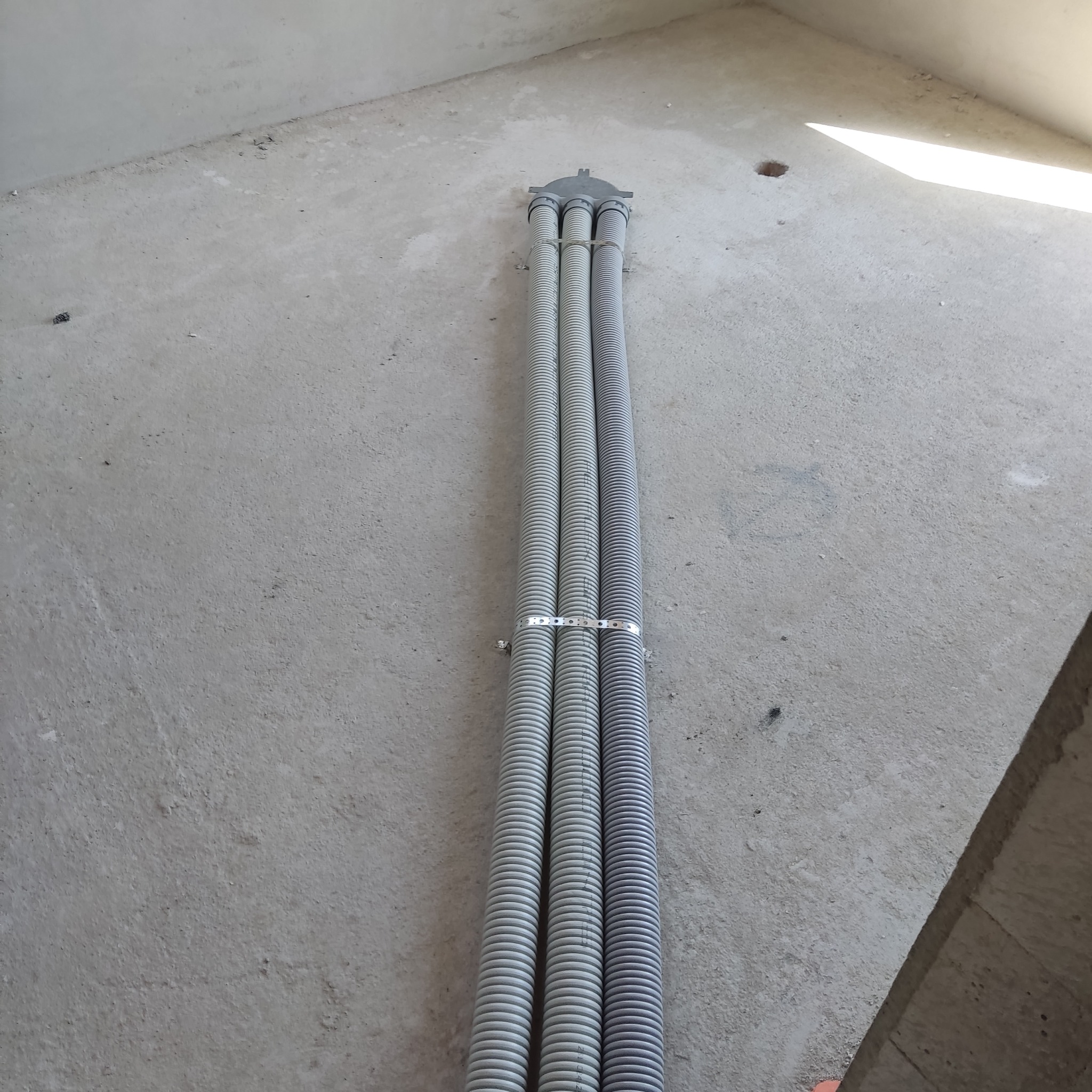 Trzy szare, karbowane rury wentylacyjne, przymocowane metalowymi obejmami do surowej, betonowej ściany w trakcie montażu.