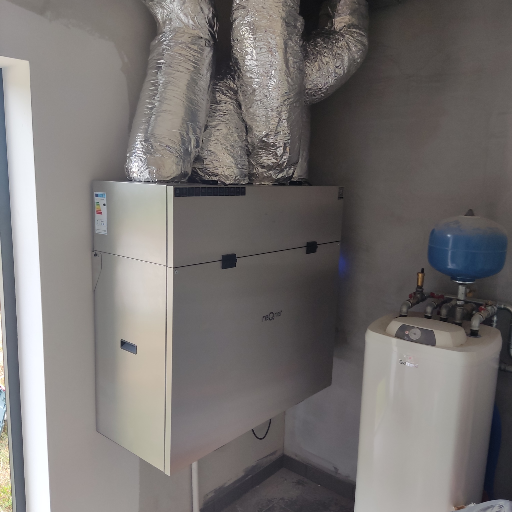 Szare urządzenie z napisem 'reQnet' połączone z srebrnymi, izolowanymi przewodami wentylacyjnymi, obok biały bojler z niebieskim zbiornikiem wyrównawczym w surowym pomieszczeniu.
