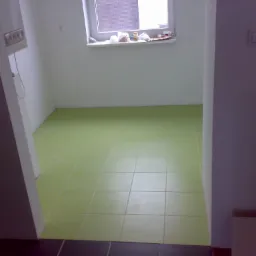 Wykończona podłoga w łazience wyłożona jasnozielonymi płytkami, widoczne fragmenty ściany i okna z parapetem.