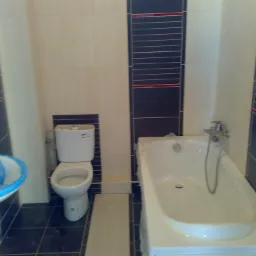 Nowa łazienka w trakcie wykańczania: biała wanna i umywalka zabezpieczone folią, toaleta oraz ciemna glazura z czerwonym akcentem.