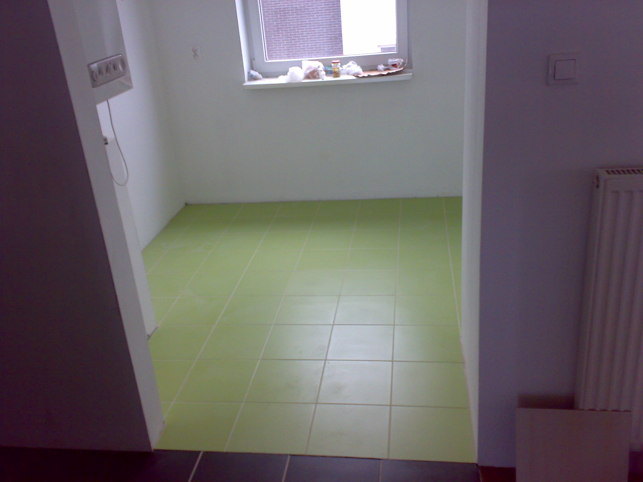 Wykończona podłoga w łazience wyłożona jasnozielonymi płytkami, widoczne fragmenty ściany i okna z parapetem.