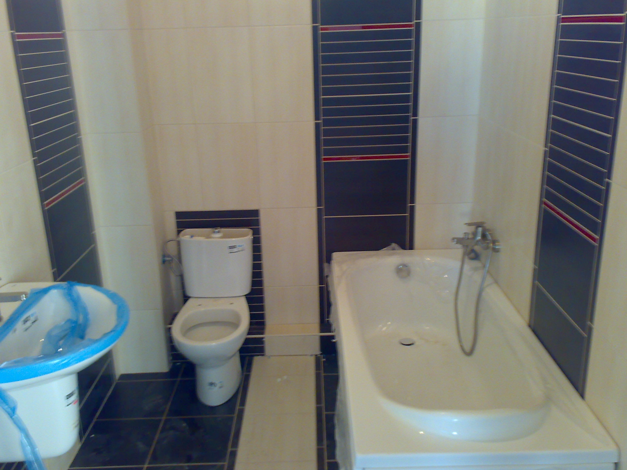 Nowa łazienka w trakcie wykańczania: biała wanna i umywalka zabezpieczone folią, toaleta oraz ciemna glazura z czerwonym akcentem.