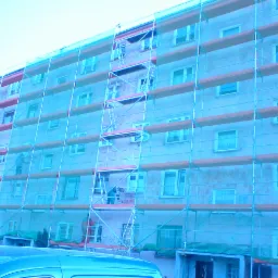 Wielopiętrowy budynek mieszkalny w trakcie prac ociepleniowych, osłonięty siatką ochronną na rusztowaniu, widoczni pracownicy na różnych poziomach rusztowania.