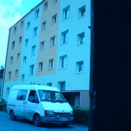 Ocieplony budynek wielorodzinny z białymi oknami, widoczny z perspektywy ulicy, z zaparkowanym białym vanem i samochodem osobowym, ujęcie częściowo zasłonięte ciemnym elementem architektonicznym.