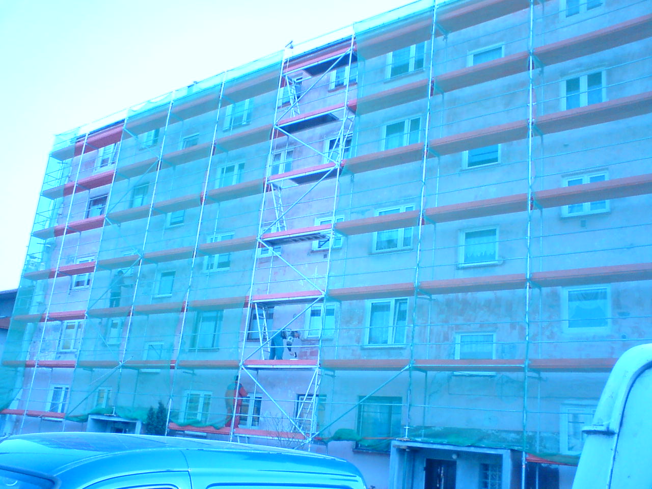 Wielopiętrowy budynek mieszkalny w trakcie prac ociepleniowych, osłonięty siatką ochronną na rusztowaniu, widoczni pracownicy na różnych poziomach rusztowania.