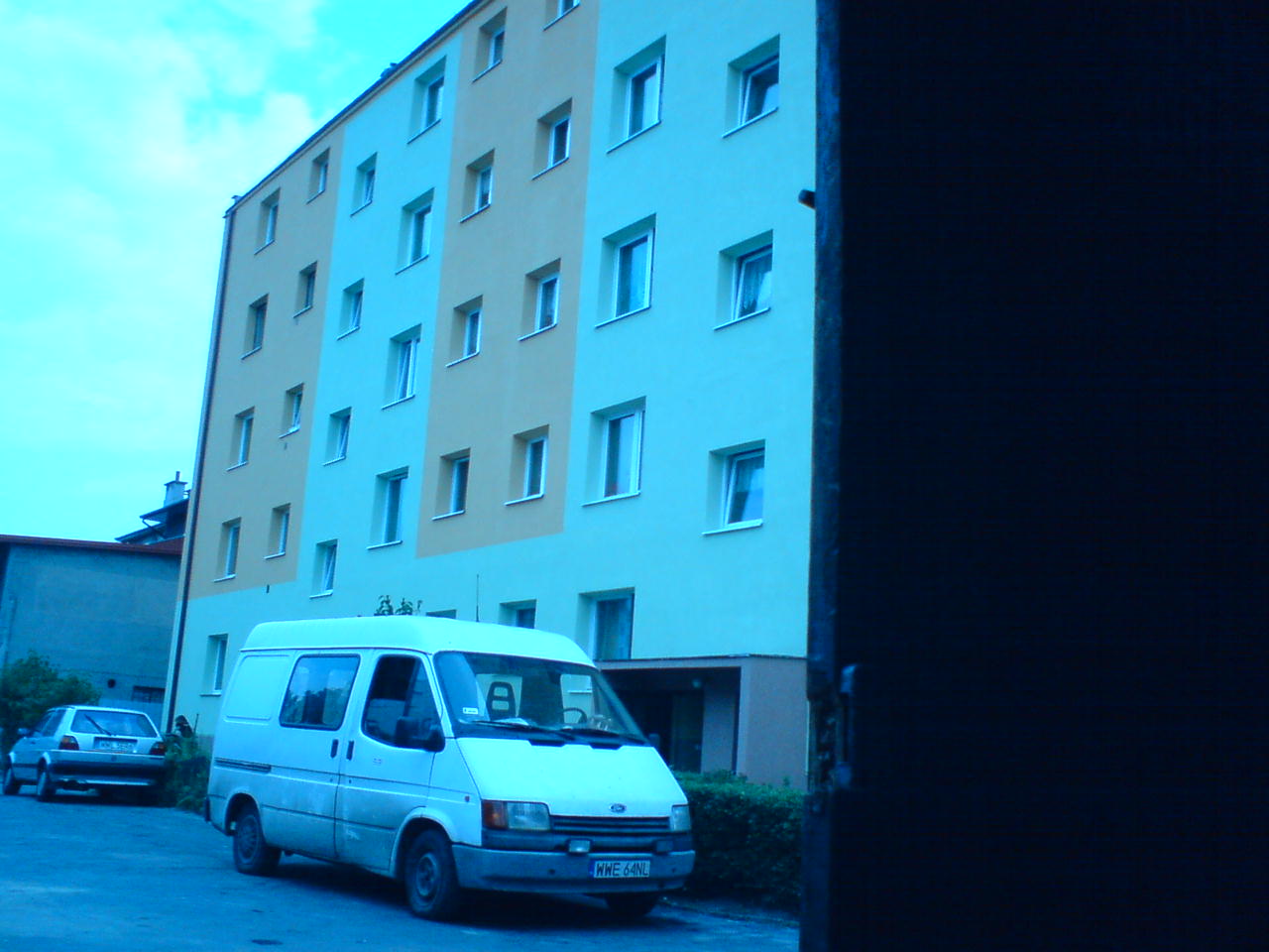 Ocieplony budynek wielorodzinny z białymi oknami, widoczny z perspektywy ulicy, z zaparkowanym białym vanem i samochodem osobowym, ujęcie częściowo zasłonięte ciemnym elementem architektonicznym.