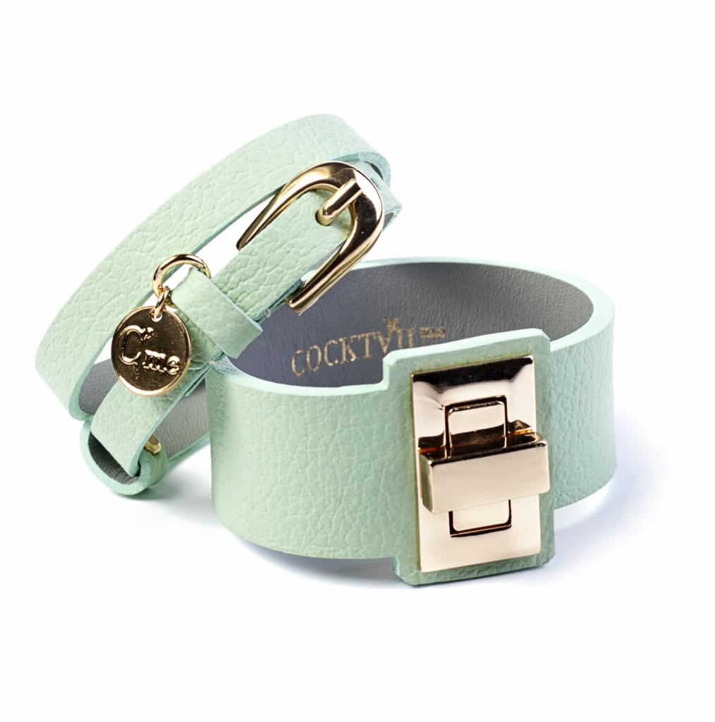 Dwa pastelowe, miętowe skórzane paski: jeden owinięty z złotą klamrą i okrągłą zawieszką z literą C, drugi szerszy z metalowym, prostokątnym zapięciem w kolorze złota, na białym tle.
