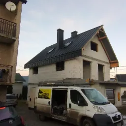 Surowy stan domu jednorodzinnego z nowym, czarnym dachem z blachy trapezowej, częściowo otynkowany, z otwartymi oknami i drzwiami, na tle błękitnego nieba, obok zaparkowany brudny bus firmy...