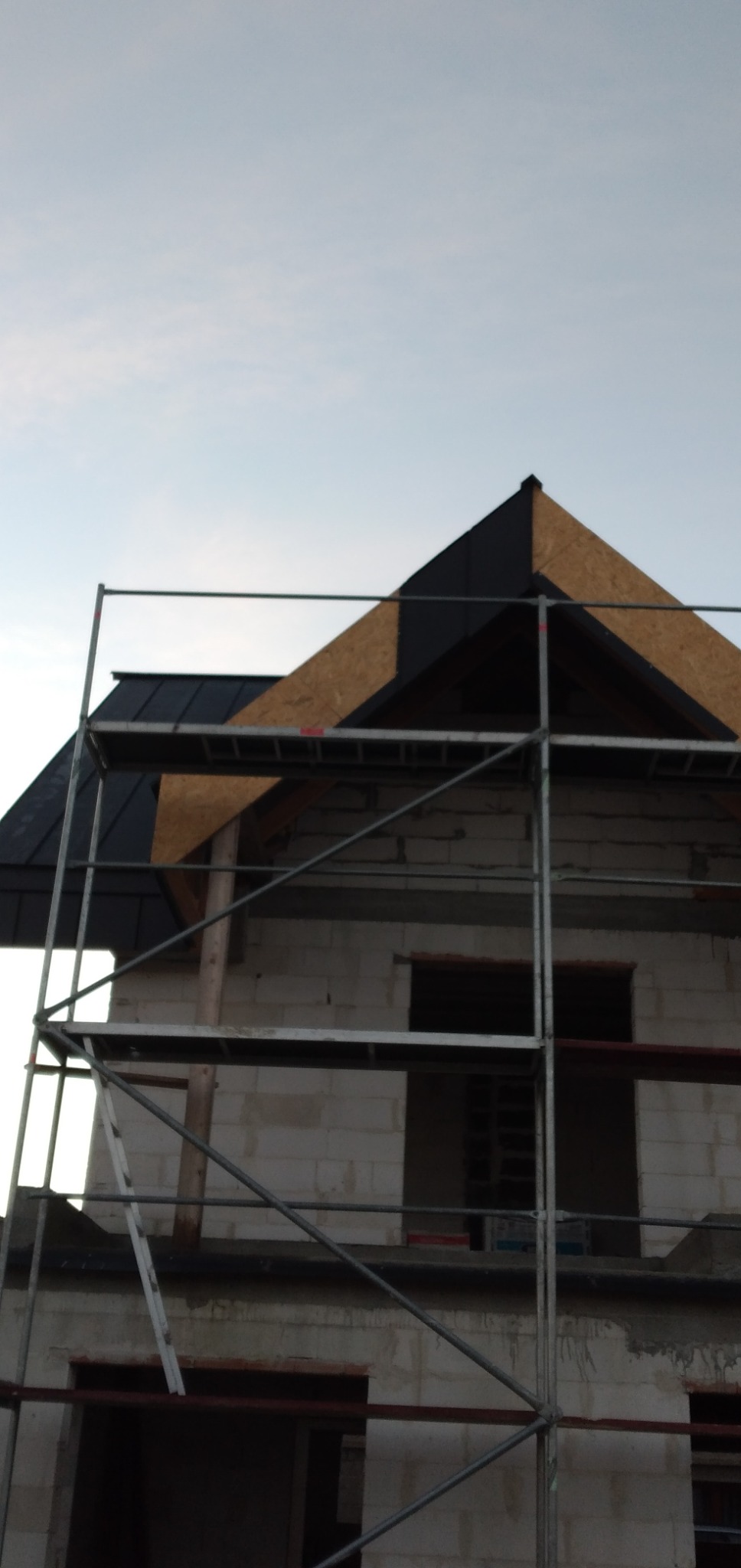 Konstrukcja domu w trakcie budowy z widocznym rusztowaniem i częściowo wykończonym dachem z płyt OSB i czarnej blachy, okna bez ram.