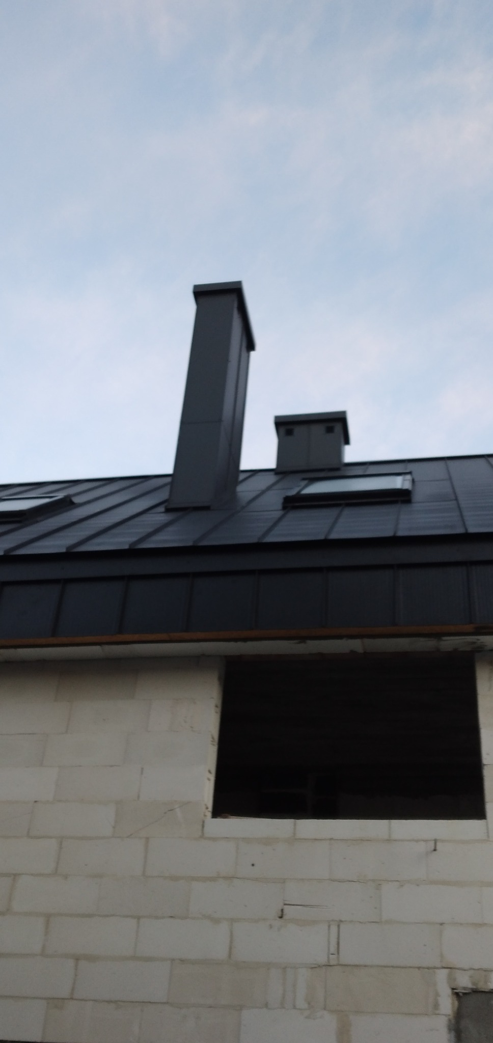 Szary dach z blachy trapezowej z dwoma kominami i oknami dachowymi, widok z dołu na budynek z pustaków, w którym brakuje okien.