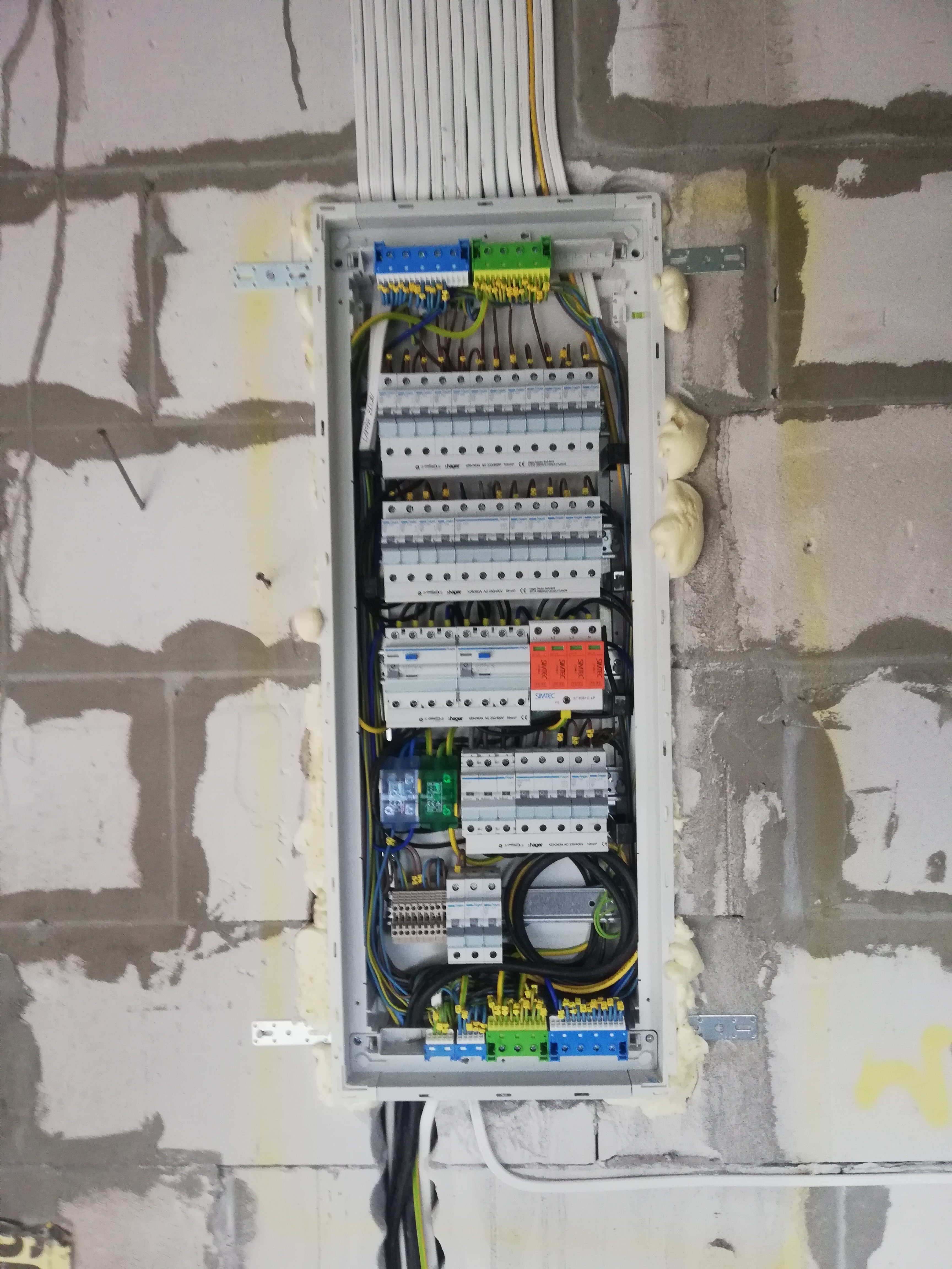 Otwarta skrzynka rozdzielcza z widocznymi bezpiecznikami, przewodami i listwami zaciskowymi, zamontowana na surowej ścianie z betonu komórkowego, uszczelniona pianką montażową.
