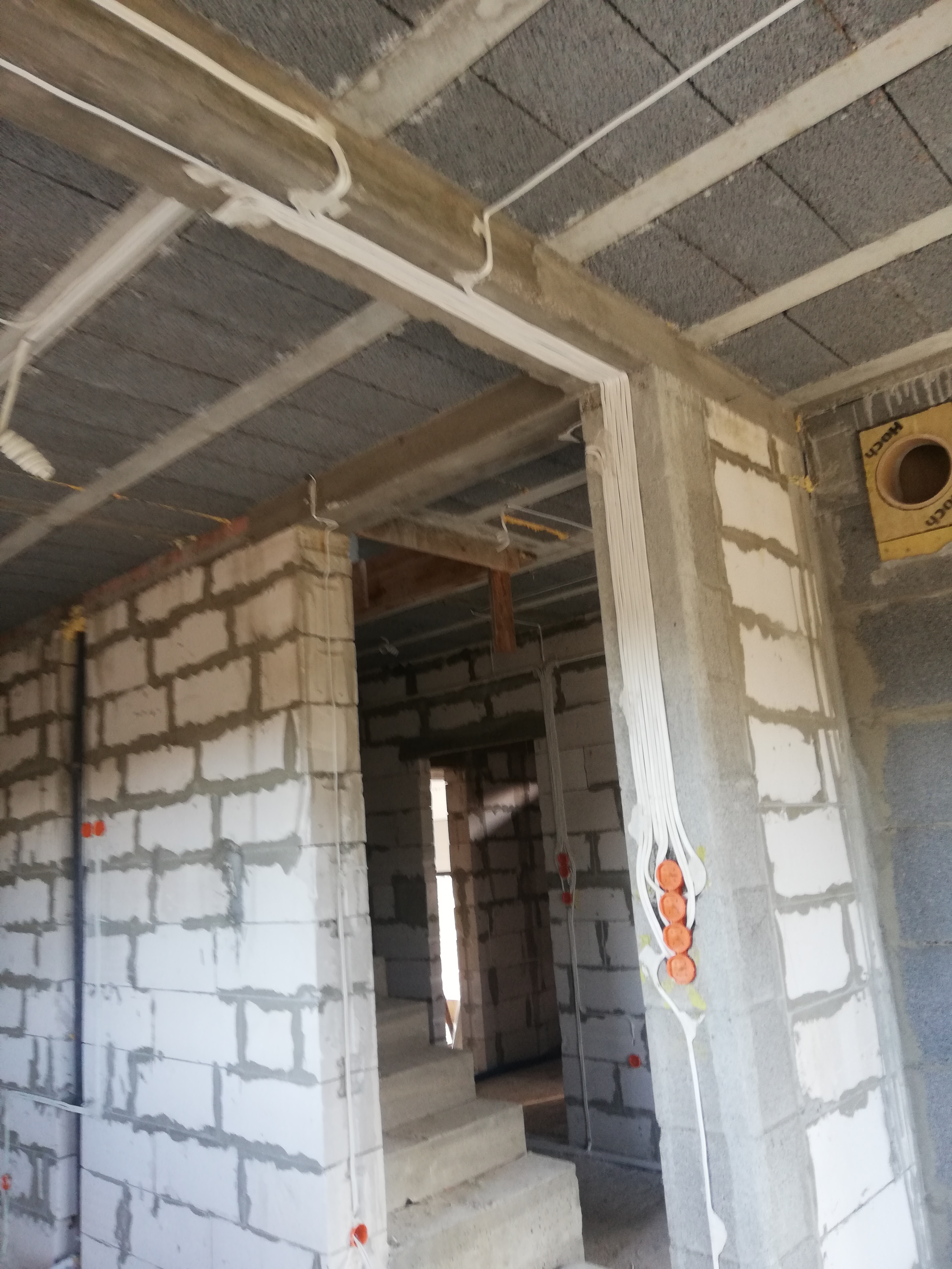 Surowy stan budynku z widocznymi, białymi kablami elektrycznymi ułożonymi wzdłuż ścian i sufitu, pomarańczowe puszki elektryczne. Szare bloczki betonowe.