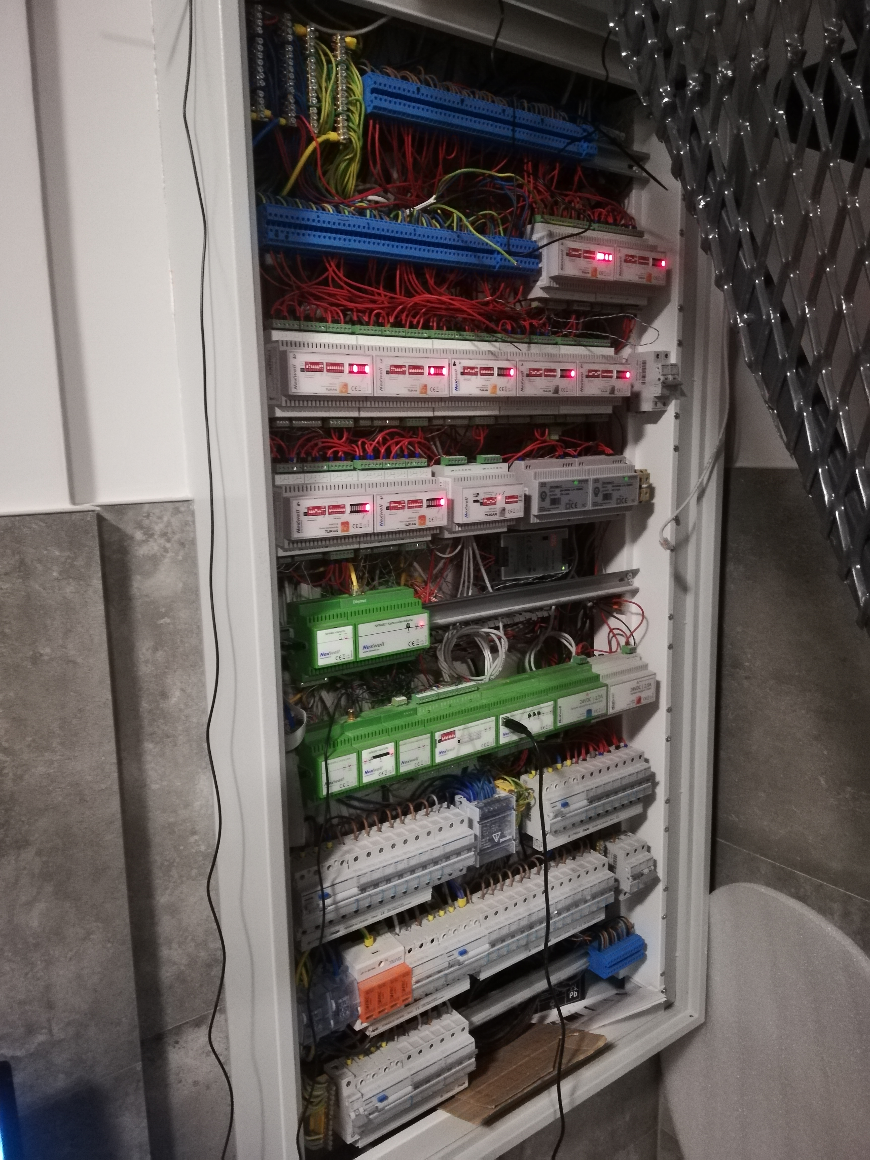 Otwarta szafa sterownicza z licznymi modułami elektronicznymi Nexwell, przewodami w różnych kolorach, listwami zaciskowymi i elementami zabezpieczającymi.