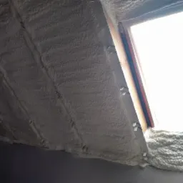 Poddasze z oknem dachowym zaizolowane białą pianą, widoczne metalowe mocowania.