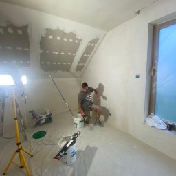 Mal-Pol - Pracownik w trakcie nakładania gładzi na ścianę poddasza, widoczne narzędzia, wiaderka z masą, lampa oświetleniowa, okno zabezpieczone folią, ściany częściowo pokryte gładzią, sufit z widocznymi...