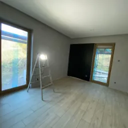 Pokój po świeżo zakończonych pracach, widoczne gładkie ściany w odcieniach szarości, drewniana podłoga, okno z widokiem na zieleń, drabina malarska z lampą halogenową i czarna ściana.