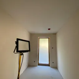 Pokój w trakcie prac przygotowawczych, podłoga zabezpieczona folią, ściany i sufit w jasnym odcieniu, widoczna lampa budowlana na statywie oraz osadzone w ścianie puszki elektryczne.