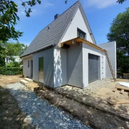 Nowoczesny dom jednorodzinny z szarą elewacją i grafitową dachówką, częściowo wykończony, z widoczną konstrukcją zadaszenia nad garażem i drabina oparta o ścianę.