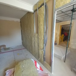 Mal-Pol - Montaż izolacji z wełny mineralnej w metalowym stelażu, widoczne przewody elektryczne i fragment pomieszczenia w trakcie prac wykończeniowych.