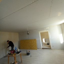 Mal-Pol - Pokój w trakcie prac wykończeniowych: szpachlowanie ścian, widoczne płyty kartonowo-gipsowe na suficie, lampa robocza, stół z narzędziami i materiałami budowlanymi, podłoga zabezpieczona przed...