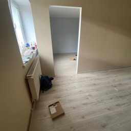 Mal-Pol - Wnętrze pokoju po malowaniu ścian na beżowo, z widoczną ościeżnicą drzwiową prowadzącą do drugiego pomieszczenia z szarą ścianą, na podłodze panele, obok grzejnika plecak i kartonowe pudełko.