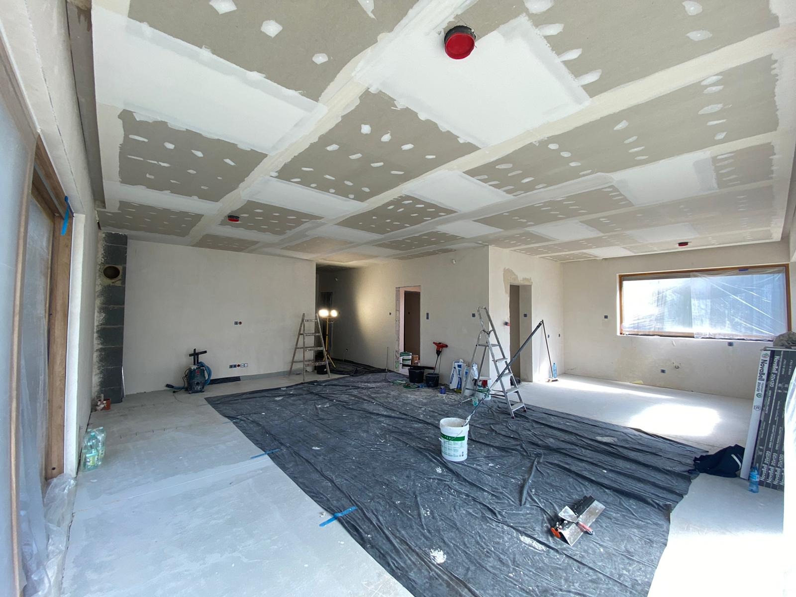 Wnętrze w trakcie prac wykończeniowych: sufit z płyt kartonowo-gipsowych po szpachlowaniu, przygotowany do malowania; podłoga zabezpieczona folią, widoczne drabiny, narzędzia i materiały budowlane.