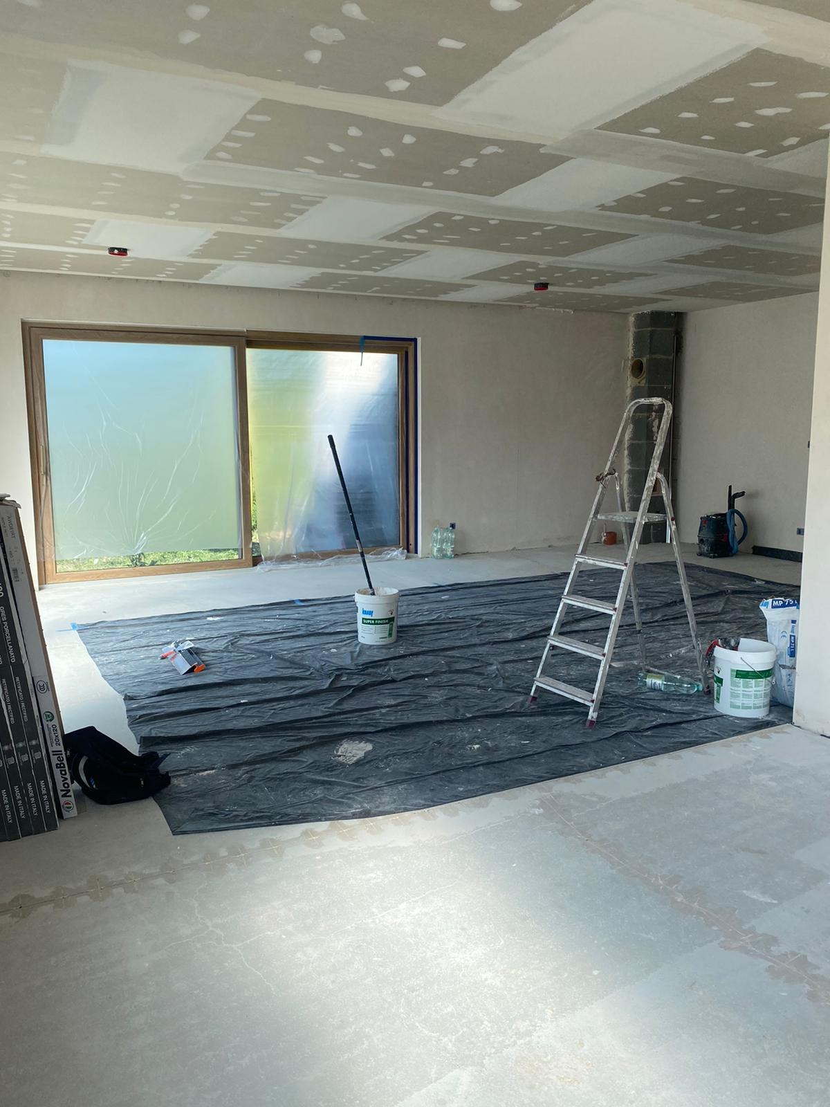 Wnętrze w trakcie prac wykończeniowych: sufit z płyt kartonowo-gipsowych po szpachlowaniu, podłoga zabezpieczona folią, drabina oparta o ścianę, widoczne wiadra z masą szpachlową i narzędzia.