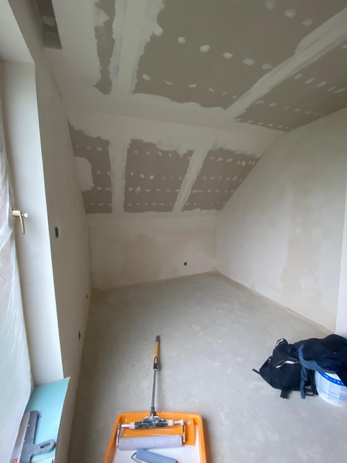 Poddasze w trakcie szpachlowania i malowania, widoczne płyty kartonowo-gipsowe na skośnym suficie, narzędzia malarskie i torba robocza na podłodze.
