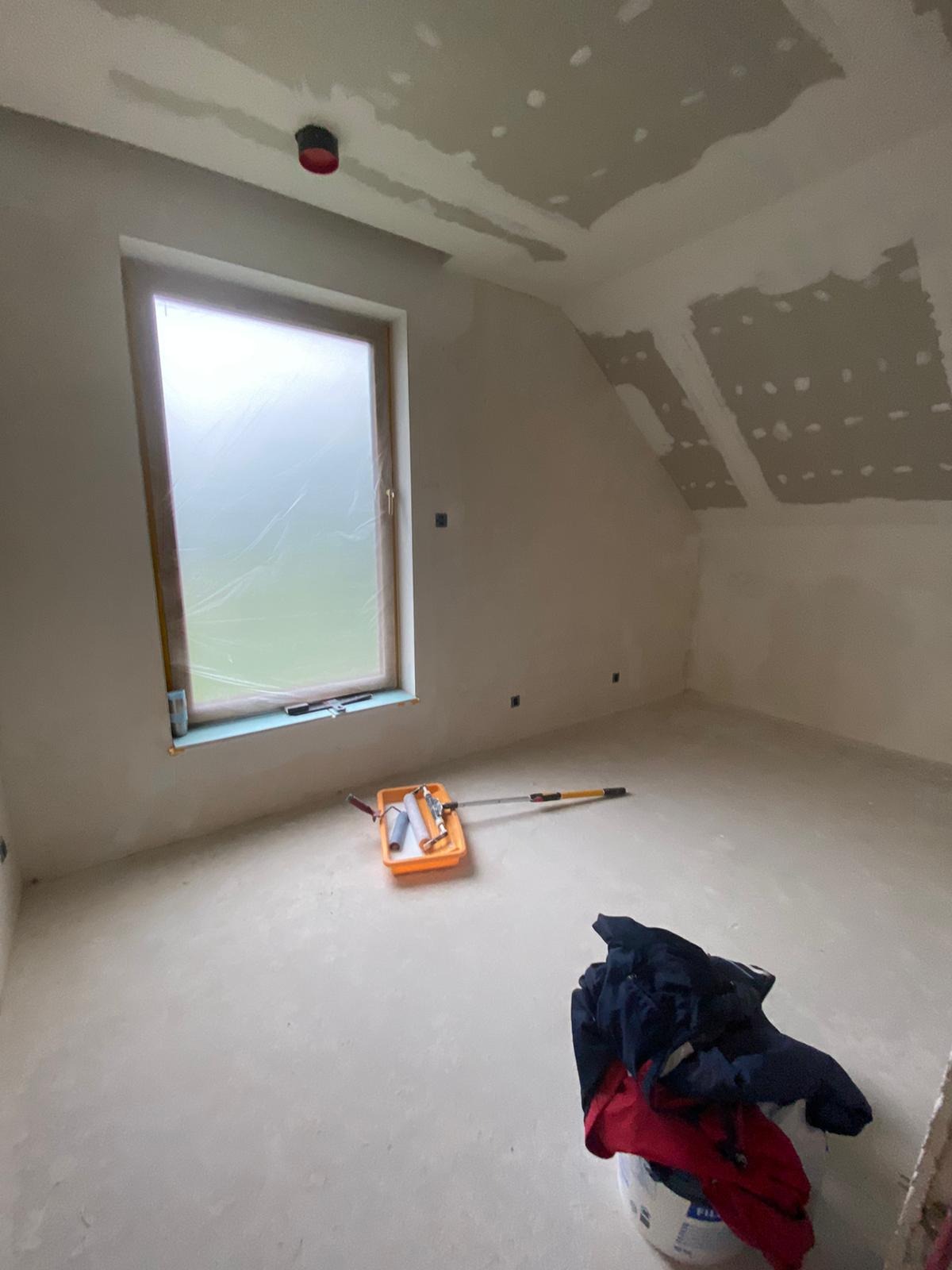 Pokój na poddaszu w trakcie przygotowań do malowania, okno zabezpieczone folią, widoczna kuweta z wałkami, ściany wstępnie wygładzone.